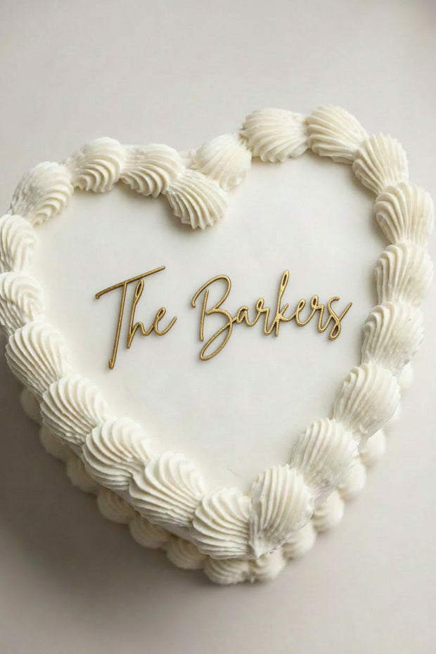 Wedding cake toppers personalized acrylic last name sign on heart shaped wedding cake with piped white frosting on soft light background