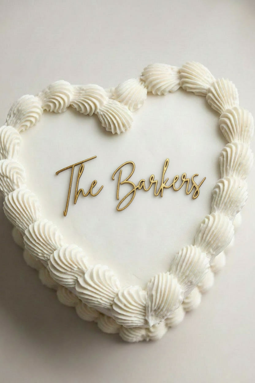 Wedding cake toppers personalized acrylic last name sign “The Barkers” on white heart shaped wedding cake with piped frosting on light background

